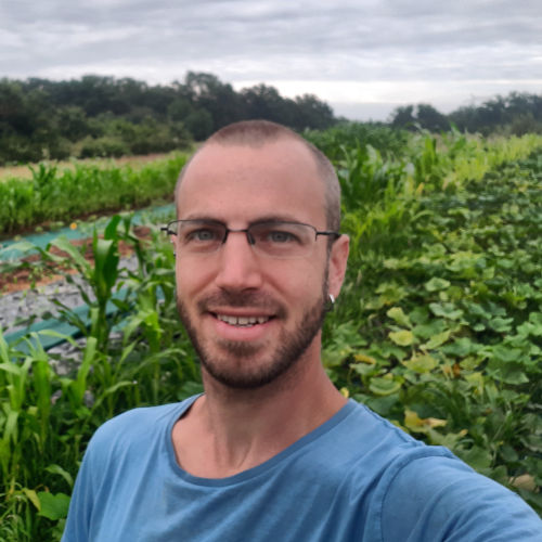 photo en buste d'un homme souriant devant un champ de milpa (courges, haricots et maïs)